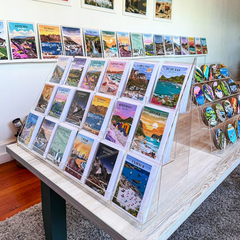 Collection of travel greeting cards on a table with additional South African travel posters displayed on the wall.