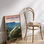 A framed travel-style poster of Blyde River Canyon, South Africa, leaning against a white wall on a light wood floor next to a curved wooden chair with a stack of books.