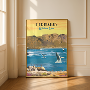 A framed vintage-style travel poster of Hermanus, Western Cape, leaning against a cream-colored paneled wall on a wooden floor. The poster features a golden-yellow sky, and blue ocean with whales