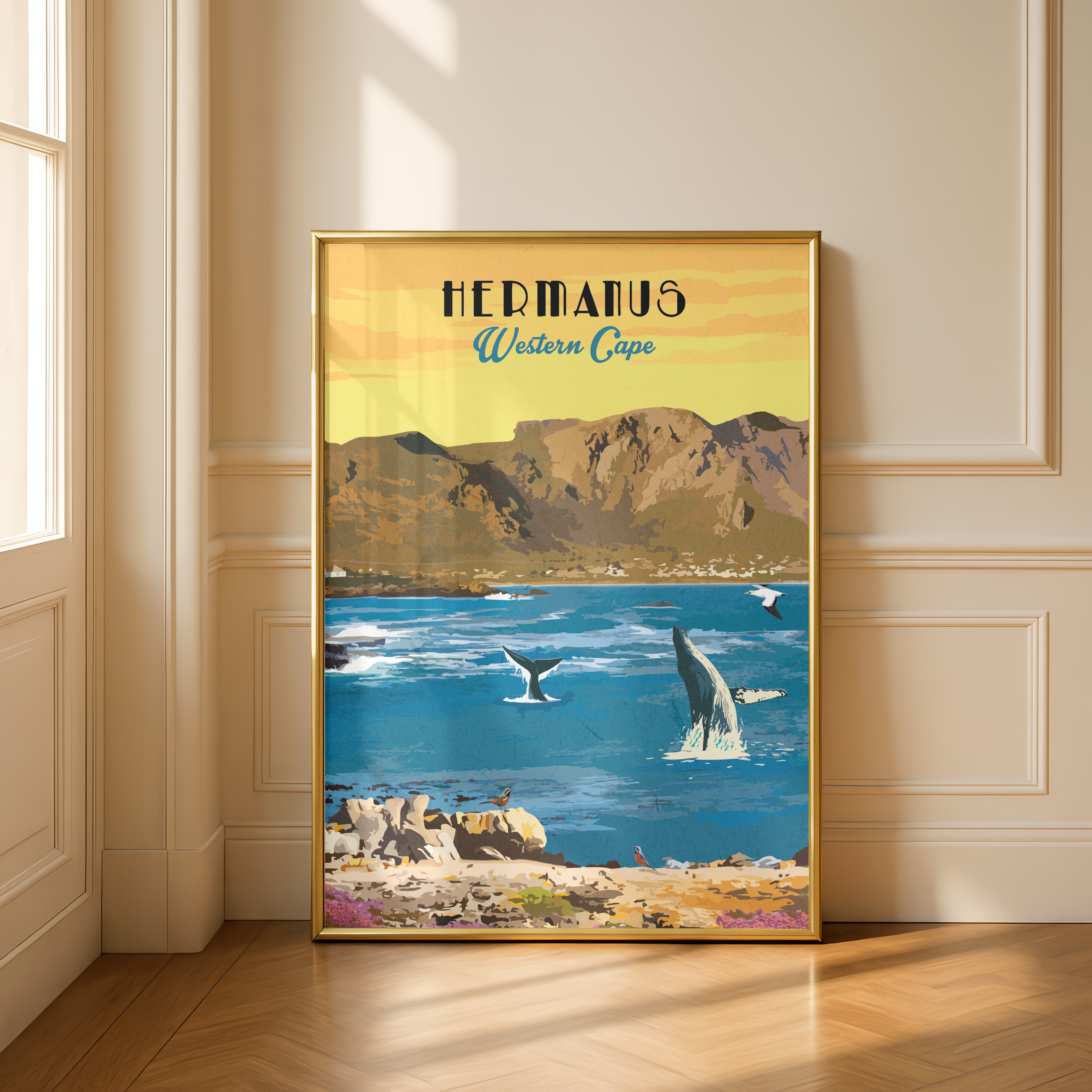 A framed vintage-style travel poster of Hermanus, Western Cape, leaning against a cream-colored paneled wall on a wooden floor. The poster features a golden-yellow sky, and blue ocean with whales