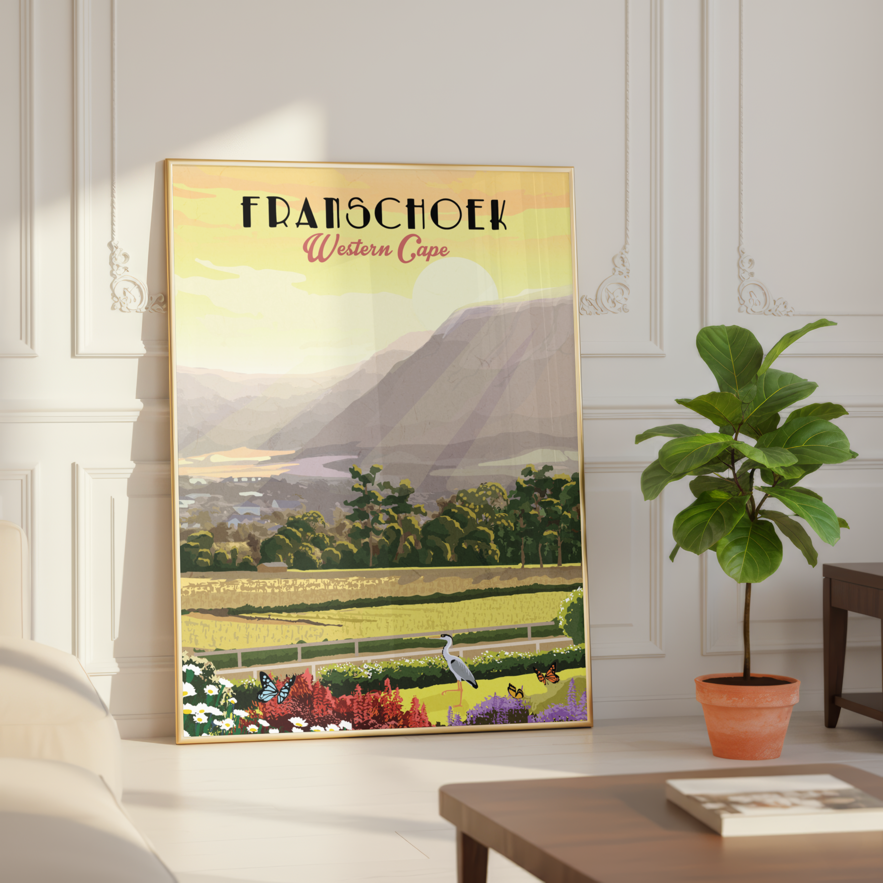 A framed vintage-style travel poster of Franschoek, Western Cape, leaning against a white paneled wall in a styled room next to a potted fiddle-leaf fig plant.