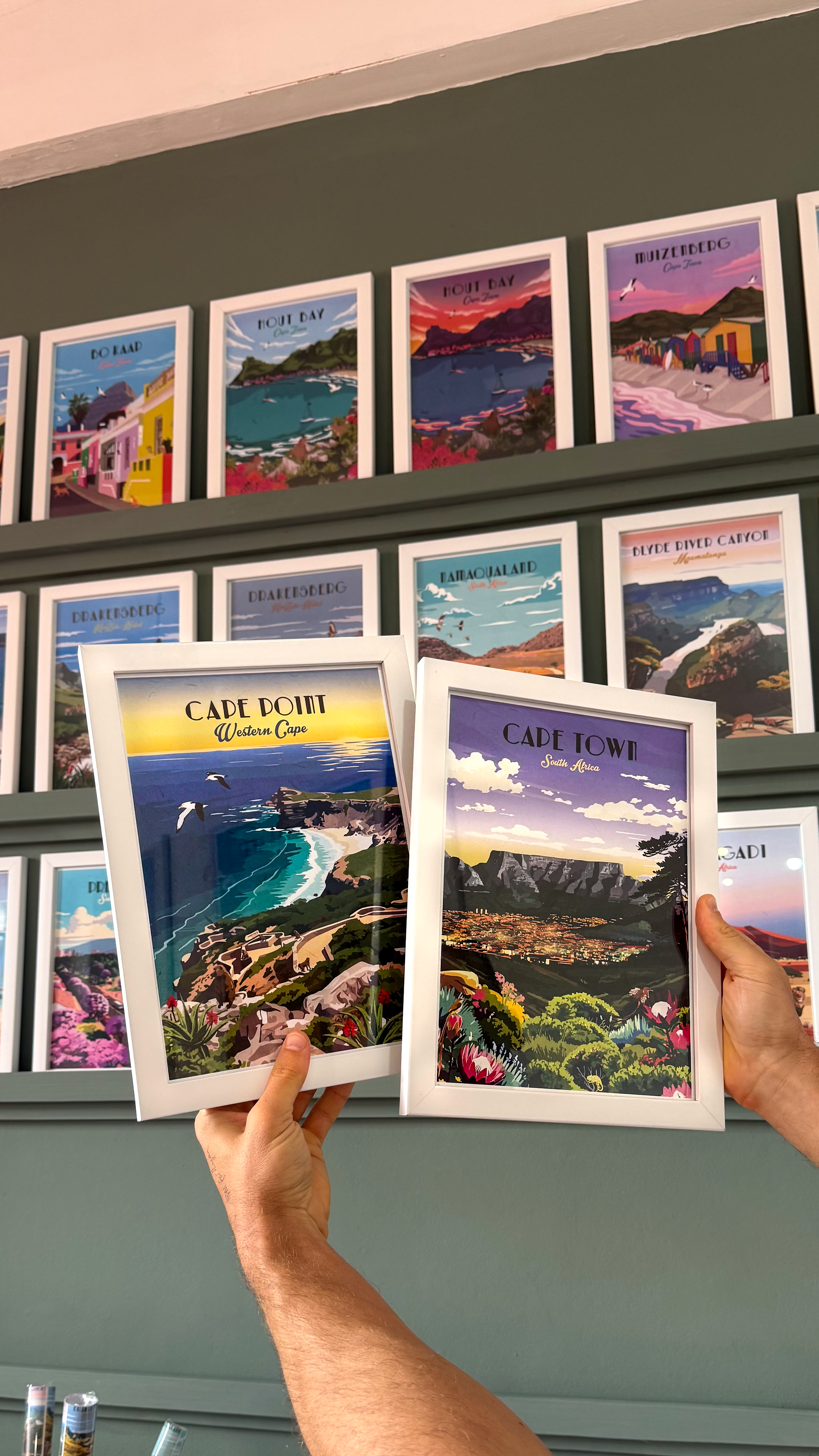Person holding a framed print of a scenic landscape next to other framed prints on a wall.