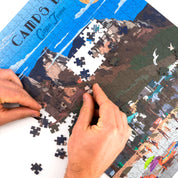 Person assembling a puzzle with a scenic design on a white surface
