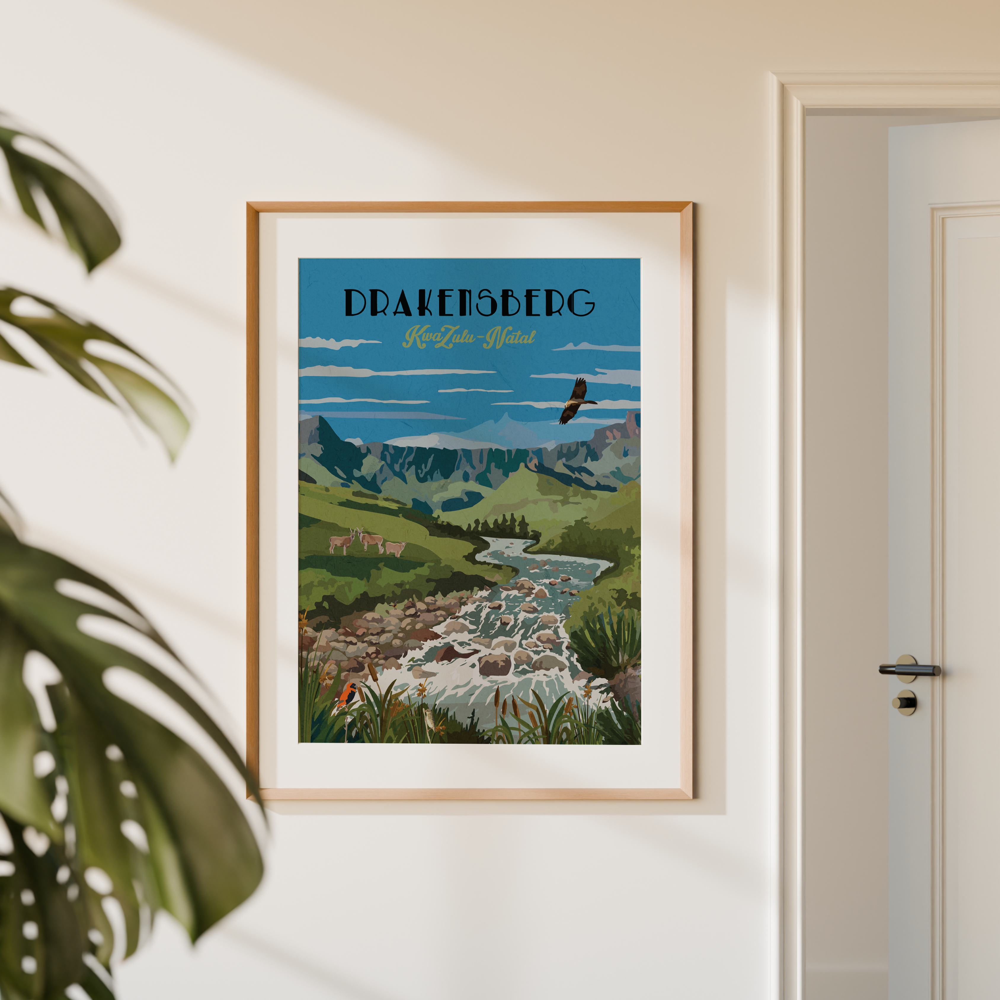 A framed travel poster of Drakenberg, styled on a warm beige wall next to a door and a green plant leaf. The poster features a vintage-style illustration of a mountain valley with a river