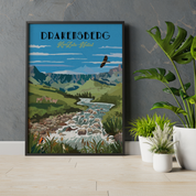 A framed travel poster of Drakensberg, South Africa, leaning against a textured gray wall on a light wood floor next to potted green plants.