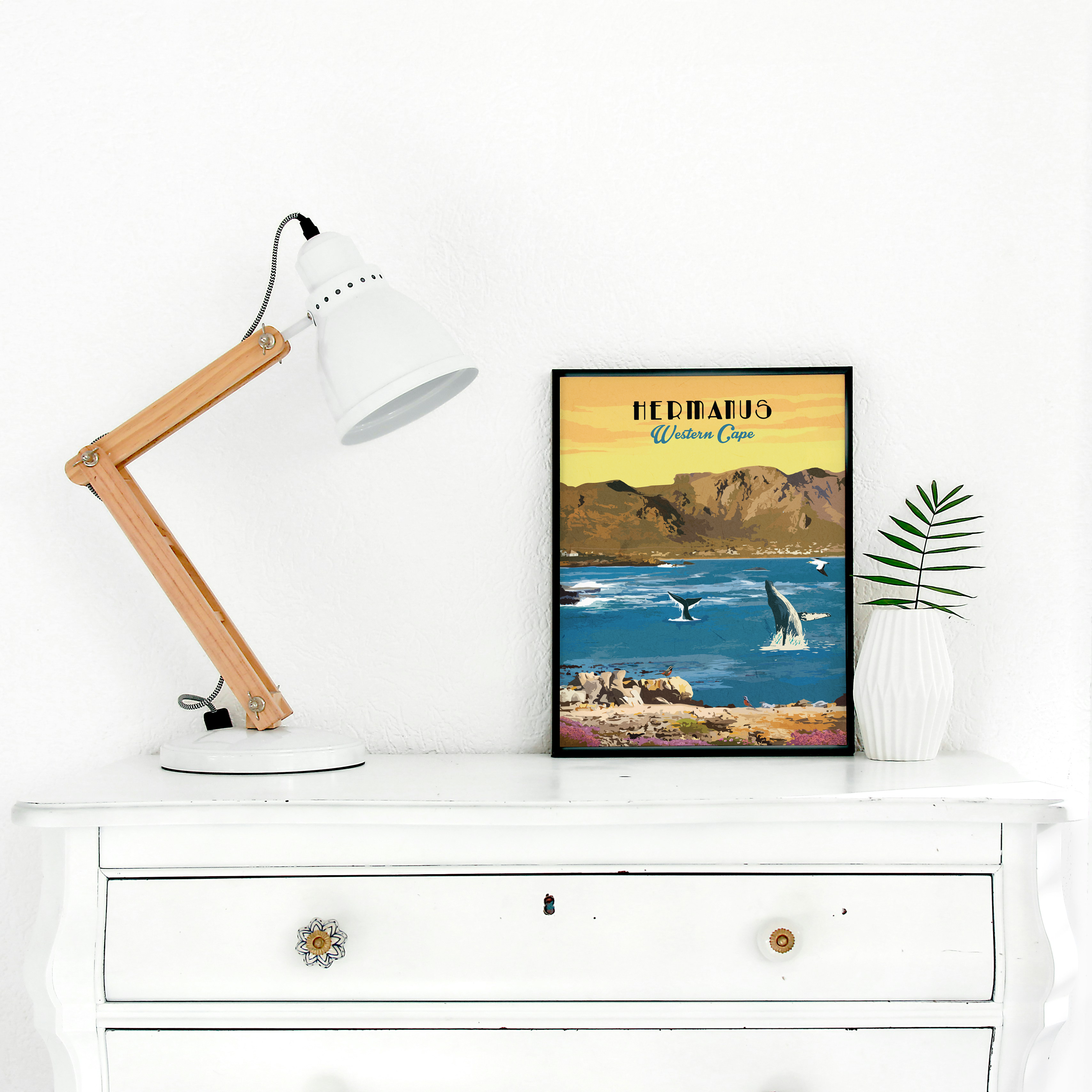 A framed vintage-style travel poster of Hermanus, Western Cape, displayed on a white dresser next to a wooden desk lamp and a small potted plant.