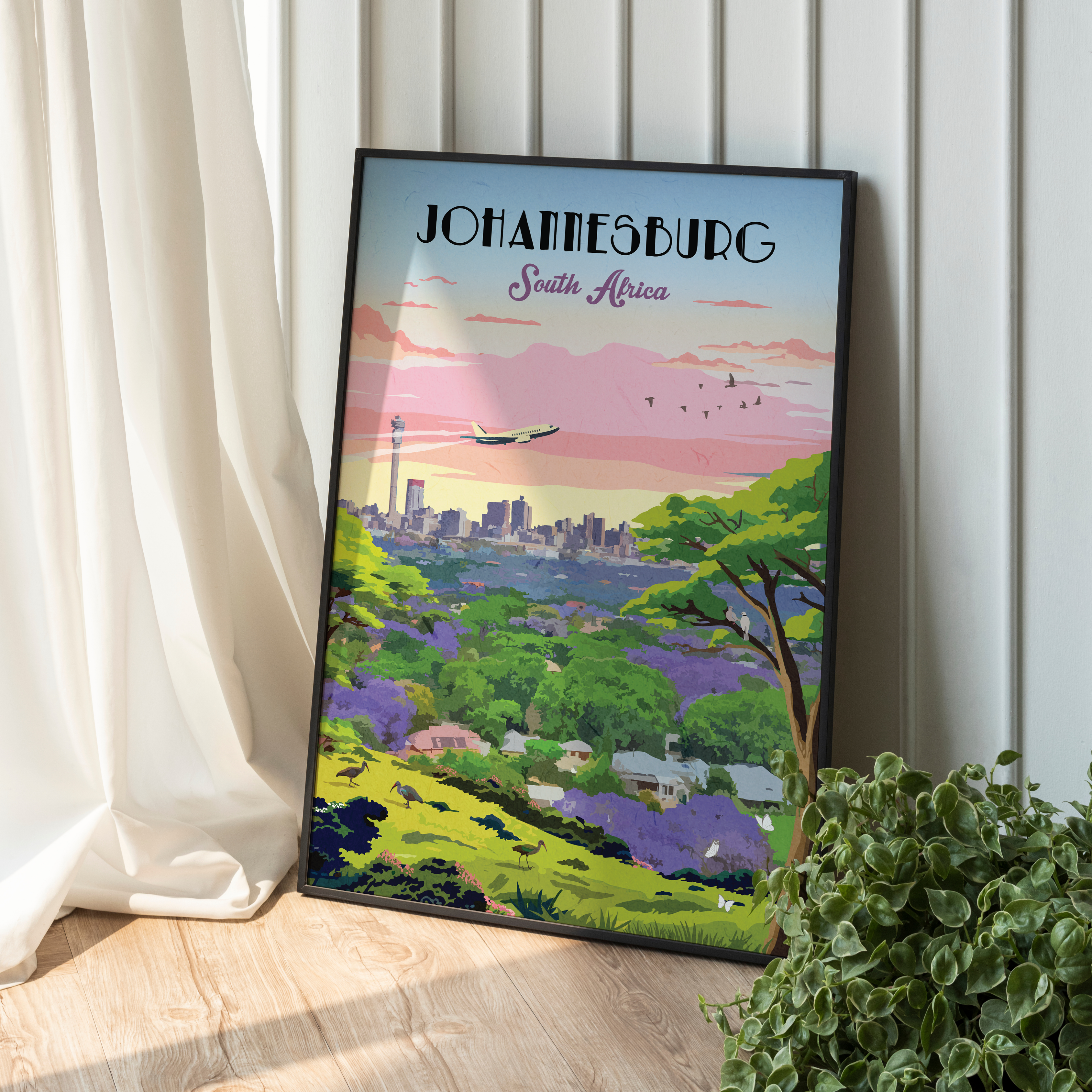 A framed travel poster of Johannesburg, South Africa, leaning against a white paneled wall on a light wood floor next to a white curtain and a green potted plant. The poster features a colorful retro illustration of a cityscape, mountains, and a  pink and blue sky.
