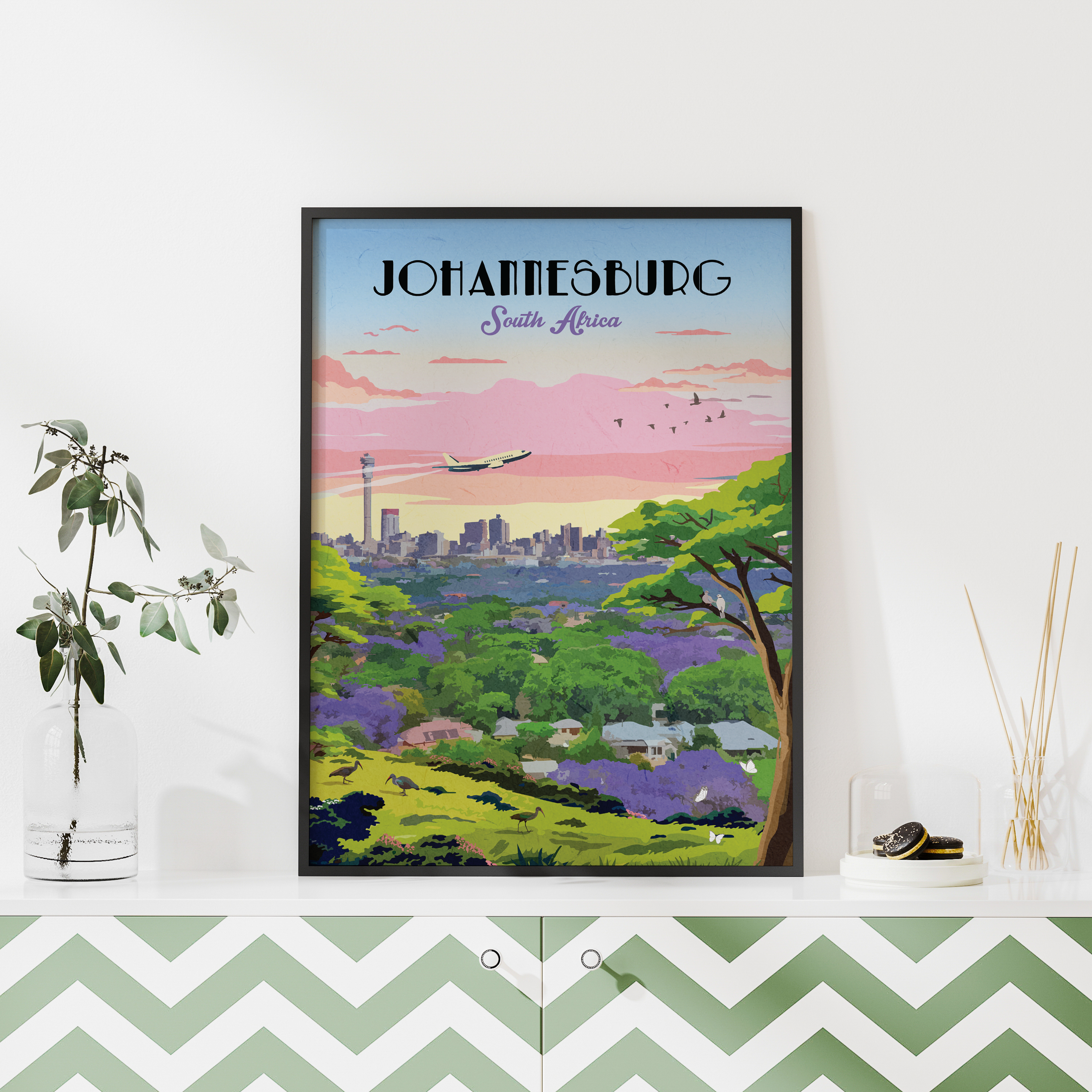 A framed travel poster of Johannesburg, South Africa, styled on a white shelf next to a glass vase with green leaves and a reed diffuser, against a white wall.