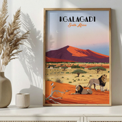 A framed vintage-style travel poster of the Galagadi dune in South Africa, featuring lions in a desert landscape, displayed on a white shelf next to a vase with dried grass.