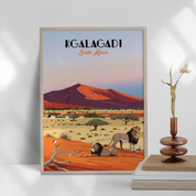 A framed travel poster of Kalagadi, South Africa, styled on a shelf next to a wooden vase with dried branches. The poster features a retro illustration of a desert landscape with red sand dunes, lions, acacia trees, and a blue sky.