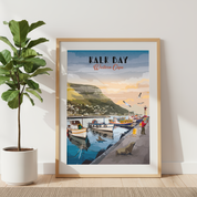 A framed art print of 'Kalk Bay Western Cape' leaning against a white wall next to a potted fiddle-leaf fig plant. The print features a vintage-style illustration of a coastal harbor with fishing boats, a green hillside town, and a warm sunset sky, enclosed in a light wood frame.