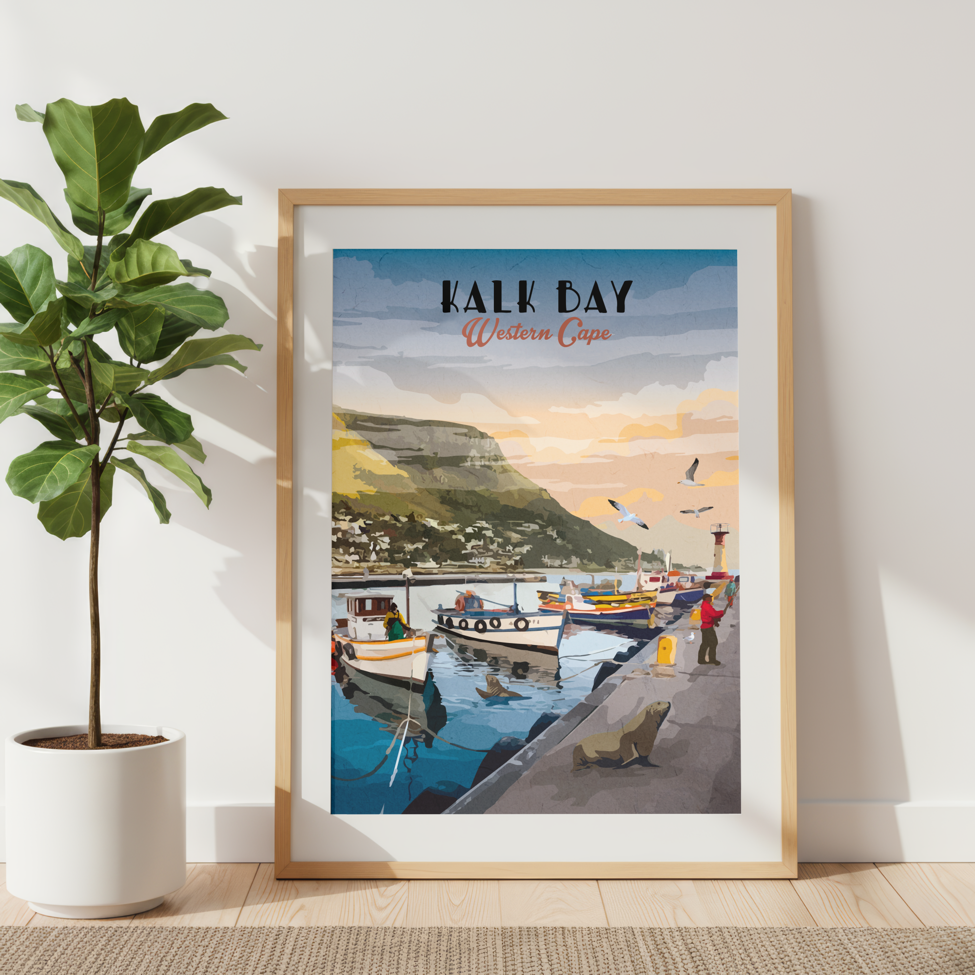 A framed art print of 'Kalk Bay Western Cape' leaning against a white wall next to a potted fiddle-leaf fig plant. The print features a vintage-style illustration of a coastal harbor with fishing boats, a green hillside town, and a warm sunset sky, enclosed in a light wood frame.