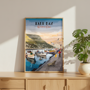 A framed art print of 'Kalk Bay Western Cape' leaning on a light wood cabinet next to a potted green plant. The print features a colorful illustration of a harbor scene with boats, a cliffside town, and a sunset sky, enclosed in a light wood frame.
