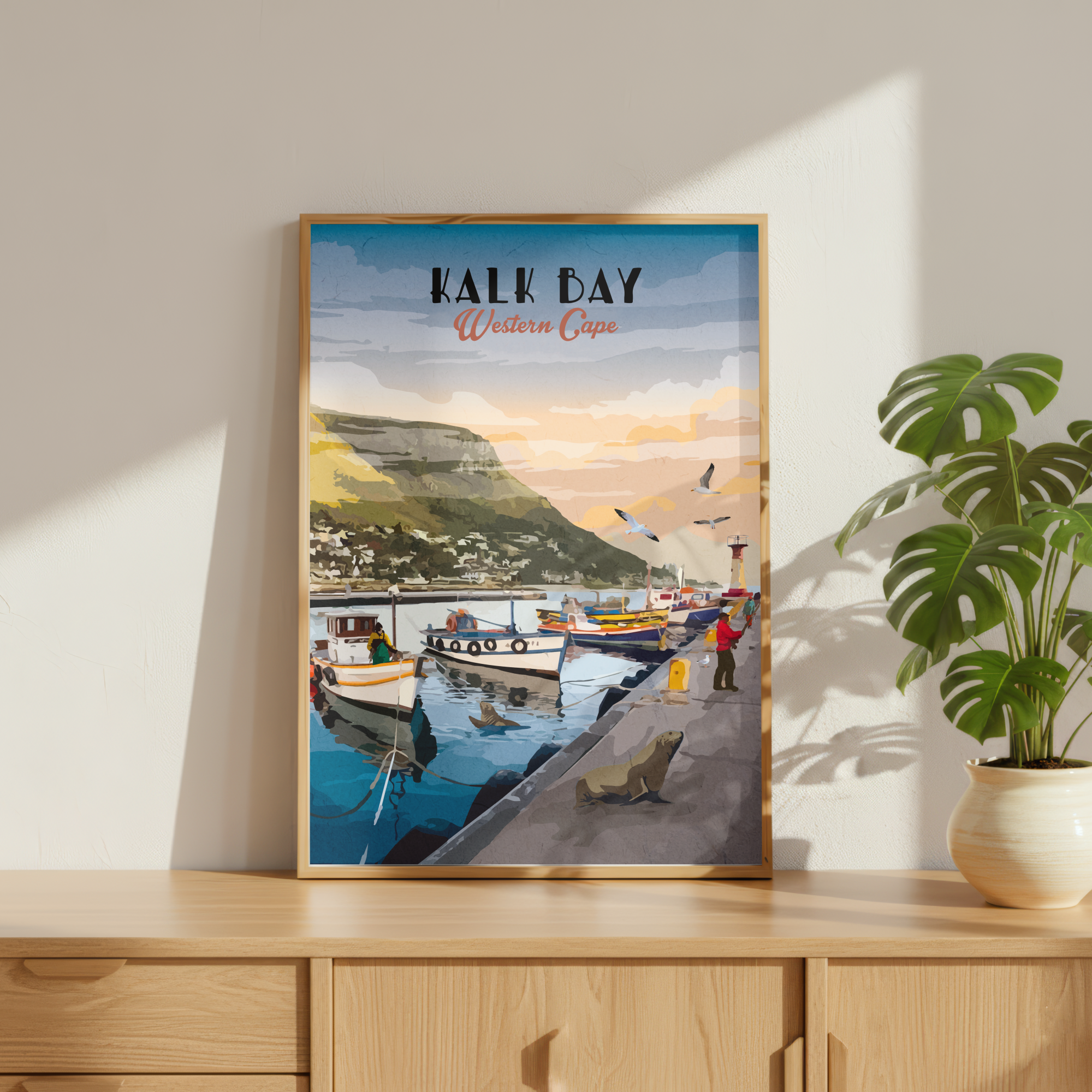 A framed art print of 'Kalk Bay Western Cape' leaning on a light wood cabinet next to a potted green plant. The print features a colorful illustration of a harbor scene with boats, a cliffside town, and a sunset sky, enclosed in a light wood frame.