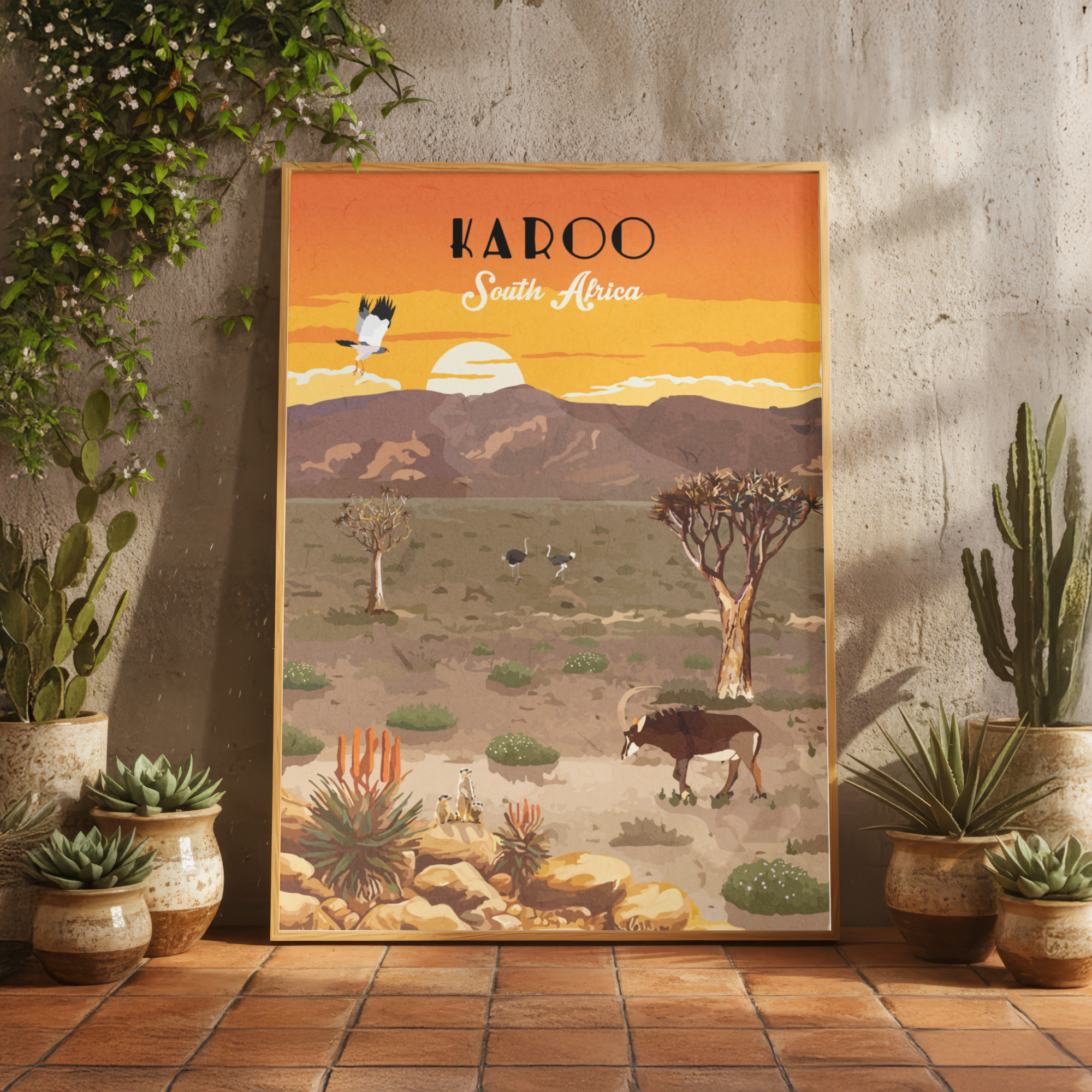 A framed art print featuring a retro-style desert landscape of South Africa with the text 'KAROO South Africa' at the top, displayed leaning against a textured concrete wall on a terracotta tile floor, surrounded by potted cacti and succulents.