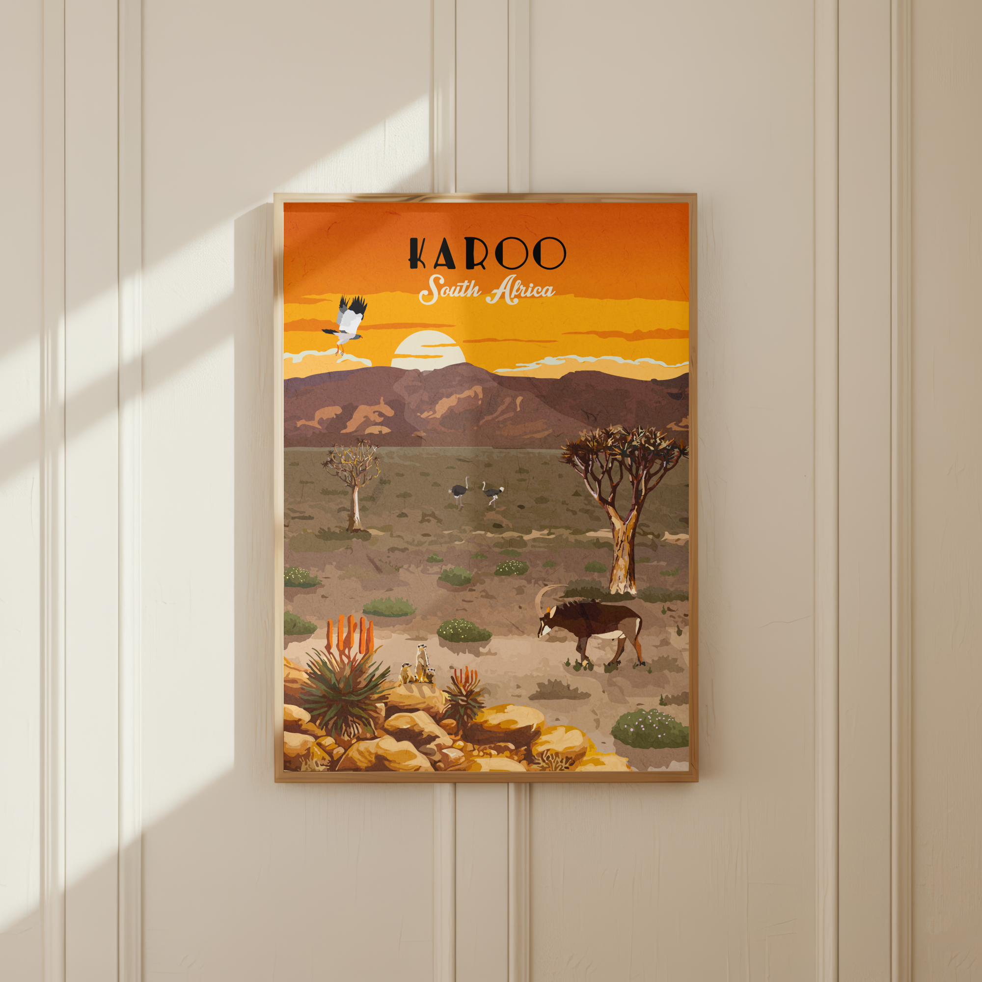 A framed travel poster of the Karoo South Africa landscape, featuring an orange sunset, mountains, and desert wildlife, displayed on a cream-colored paneled wall with soft window shadows.